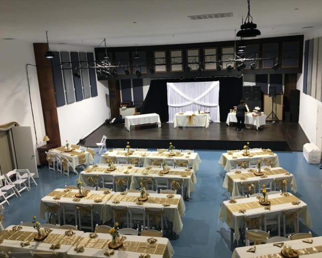 A spacious event hall is set up with neatly arranged tables covered in white cloths, each decorated with simple centerpieces, and a stage area at the front.