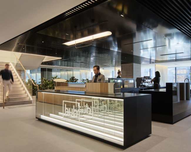 The image shows a modern office lobby with a reception desk, a staircase, and various seating areas surrounded by plants.
