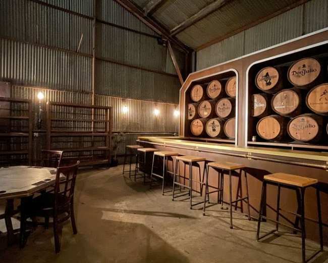 A rustic bar area inside a metal warehouse, featuring barrel-style decor and wooden furniture.