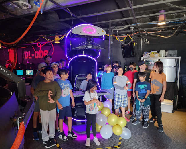 A group of children and adults stands around a brightly lit gaming chair in a dark room filled with computer stations.