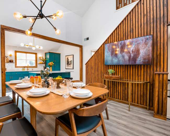 A dining area features a wooden table set for dinner, surrounded by chairs, with blue cabinets and a wall-mounted TV in the background.