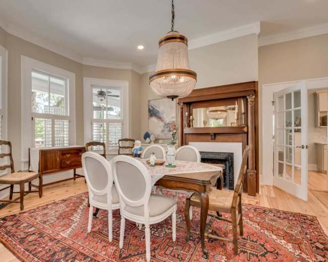 The image shows a dining room with a wooden table surrounded by chairs, a chandelier overhead, and large windows allowing natural light to enter.