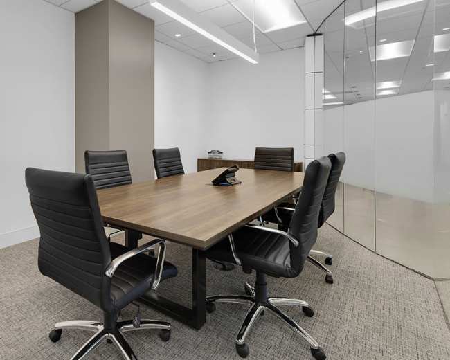 A conference room features a large wooden table surrounded by six ergonomic chairs, with a phone on the table and glass walls.