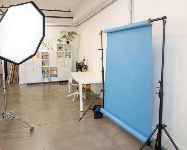 A photography studio features a blue backdrop, a large light source, and a white desk with shelves in the background.