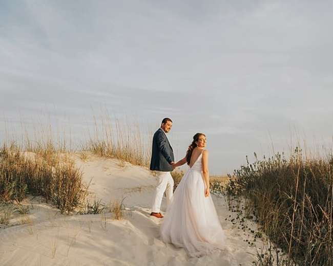 A couple walks hand in hand on a sandy dune with grass and shrubs in the background.