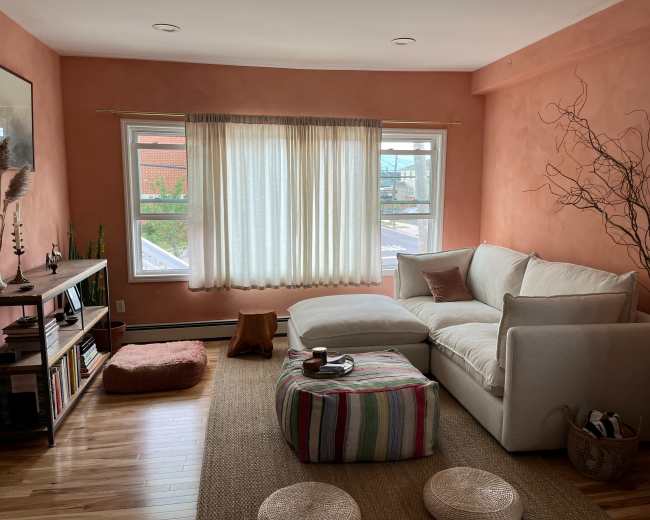 A cozy living room with a light-colored sectional sofa, a woven rug, and large windows covered by sheer curtains.