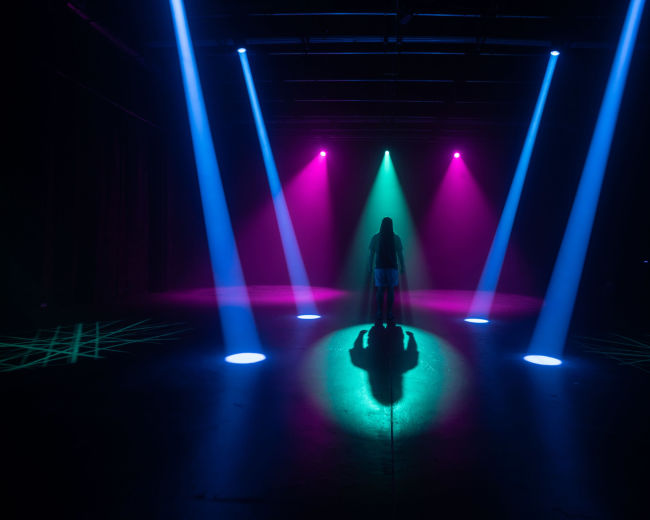 A solitary figure stands on a stage illuminated by beams of colored light, casting distinct shadows on the floor.