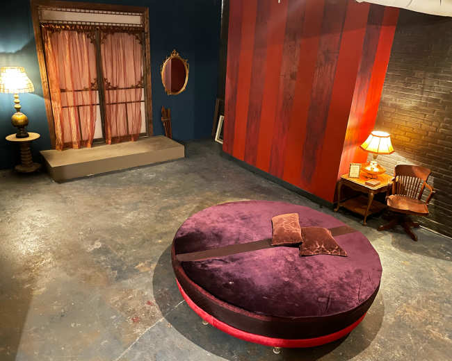 The image shows a room with a round purple seating area in the center, a red-striped wall, a vintage lamp, and a small table with a chair in one corner.