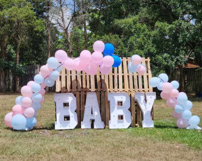 A decorative display featuring large white letters spelling "BABY" with an arch of pink and blue balloons in a grassy area.