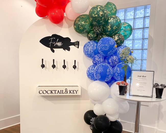 A decorative entryway featuring a white display with a fish silhouette and four drink taps, surrounded by colorful balloons and a small table with a menu.