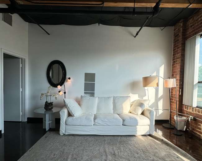 A light-colored sofa is positioned in a room featuring exposed brick walls, a large window, a floor lamp, and a mirror.