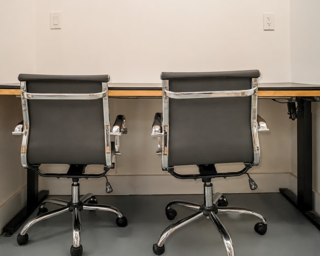 The image shows a minimalist office space with two black ergonomic chairs positioned in front of a wooden desk against a light-colored wall.