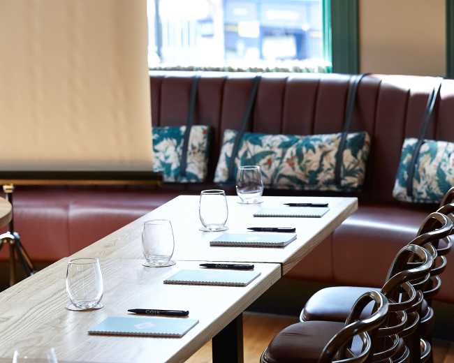 A long wooden table with place settings, glasses, and notepads is arranged in a brightly lit room with leather seating.
