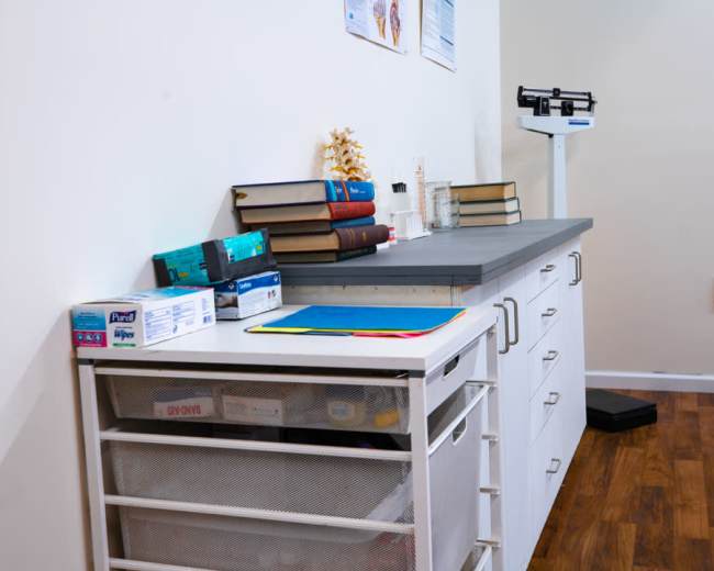 A medical examination room features a countertop with stacked books, a spine model, and medical supplies, alongside a scale and storage drawers.
