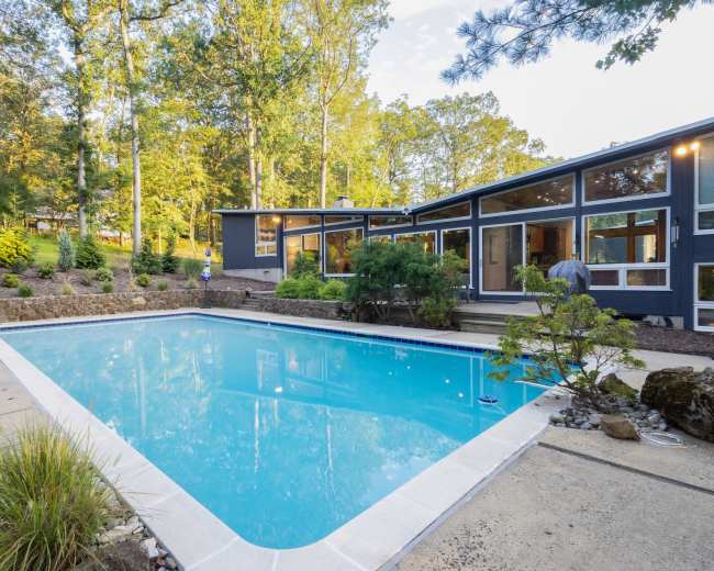 The image shows a modern home with large windows overlooking a clear swimming pool surrounded by landscaping and trees.