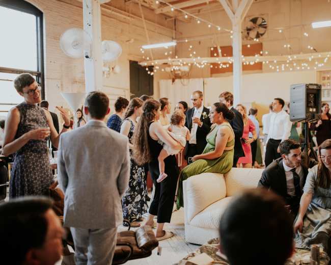 A large group of people socialize at a lively indoor event, surrounded by string lights and various seating arrangements.
