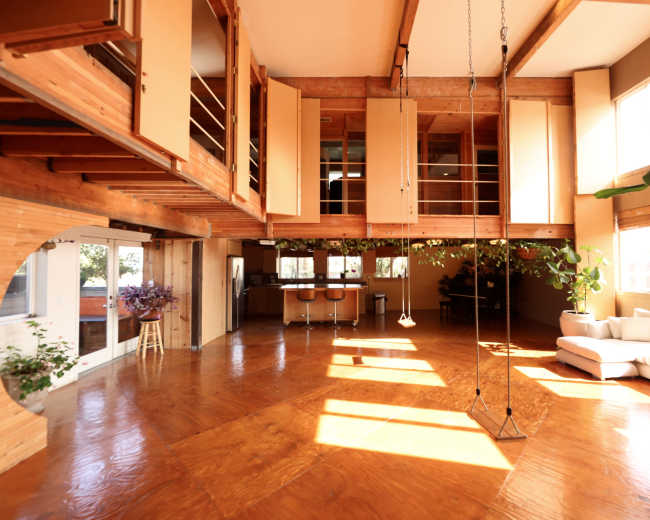 The image shows a spacious, modern interior with wooden beams, an open floor plan, a swing, and a kitchen area in the background.