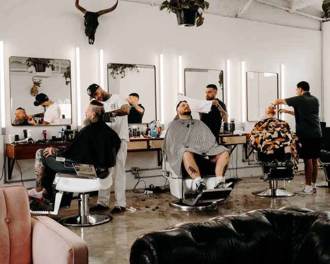 The image shows a modern barbershop with multiple clients seated in barber chairs while stylists work on their haircuts.