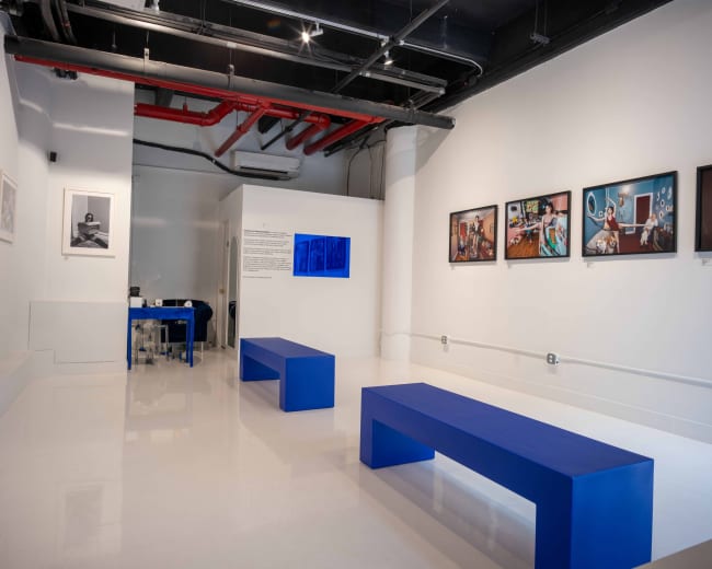 The image shows a gallery space with white walls and shiny floors, featuring blue benches and framed photographs hung on the walls.