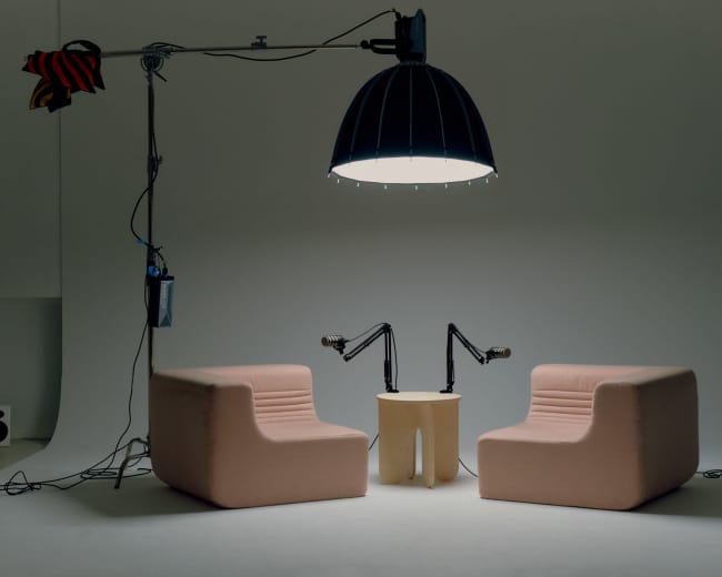 The image shows two pink, upholstered chairs facing each other, with a round wooden table between them, illuminated by a large pendant light in a minimalistic studio setting.