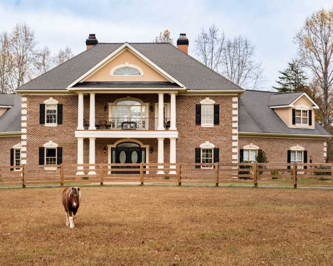 A large brick house with multiple gables and a front porch stands in a grassy field, while a brown and white cow grazes nearby.
