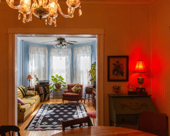 The image shows a cozy room with a living area featuring a patterned rug, comfortable seating, and large window drapes, illuminated by soft lighting.