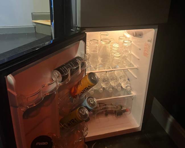 A small refrigerator with an open door displays several glass jars, water bottles, and cans on its shelves.