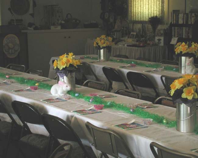 A long table is set up for a gathering, decorated with yellow flowers and small white bunny figurines.