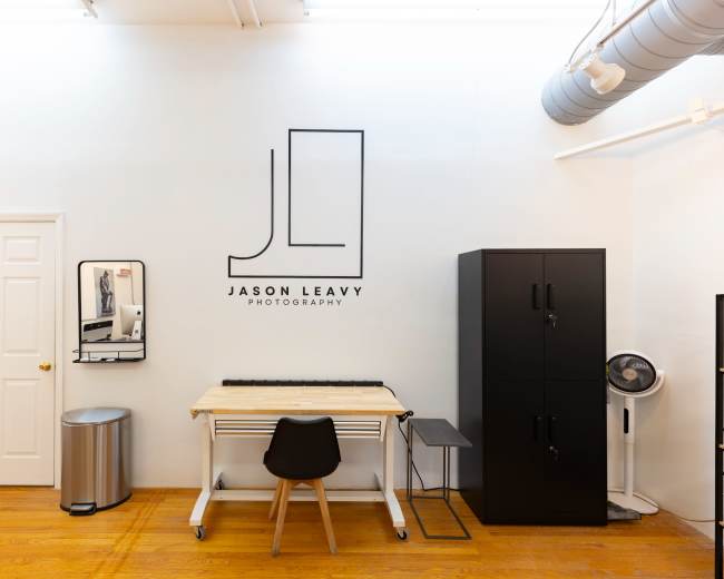 The image shows a clean photography studio with a central work table, a black cabinet, a mirror, and a door along the wall featuring the logo "JASON LEAVY PHOTOGRAPHY."