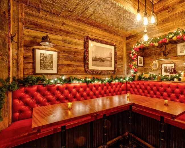 The image depicts a cozy restaurant corner with red tufted seating, wooden walls, and Christmas decorations of garlands and ornaments.