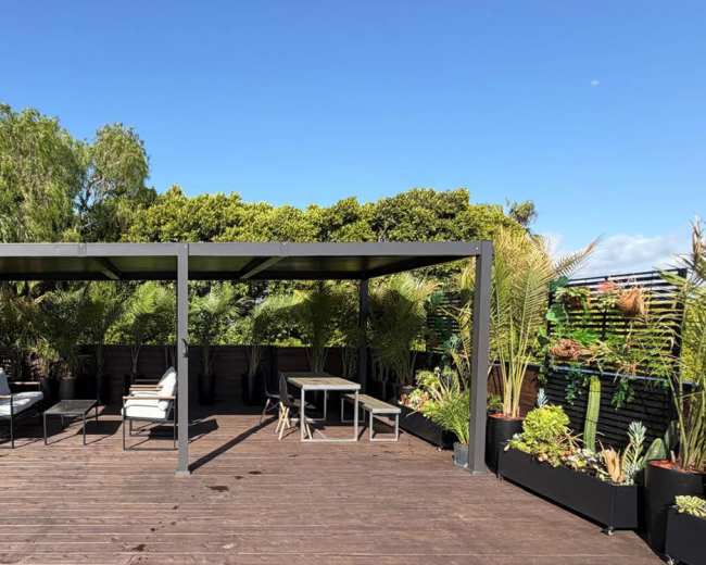 The image shows a rooftop space with a shaded metal structure surrounded by potted plants and seating areas.