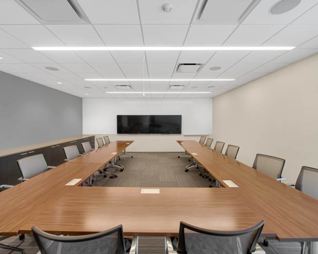 A modern conference room features a large wooden table surrounded by ergonomic chairs and an electronic display on the wall.