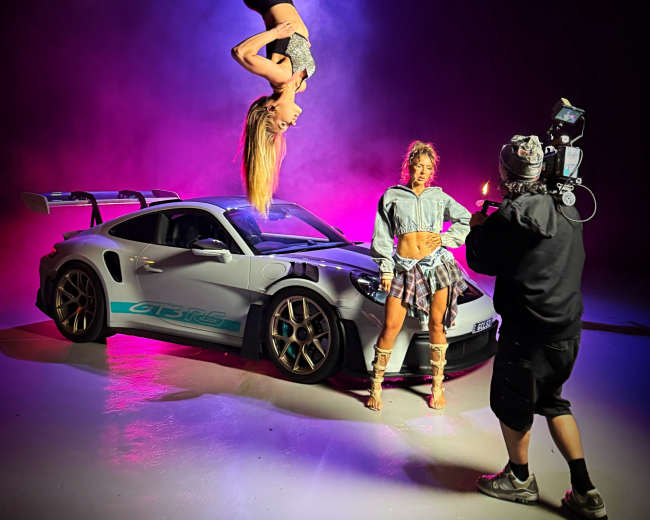 A woman performs an aerial act on a hoop above a sports car while another woman poses nearby and a photographer captures the scene.