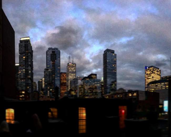 The image showcases a skyline of tall buildings and skyscrapers in a city at dusk, with a mix of illuminated windows and dark clouds overhead.