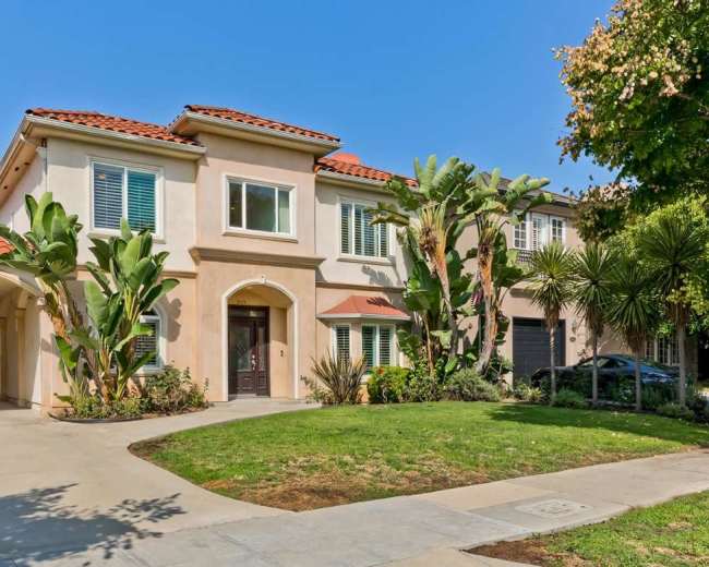 The image shows a two-story house with a tile roof, surrounded by palm trees and neatly landscaped shrubs, along a concrete walkway.