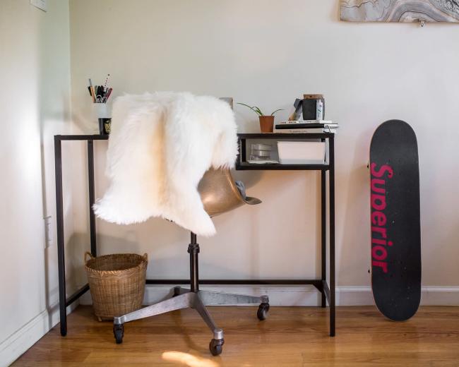 A modern workspace features a black metal desk with an ergonomic chair, a fur throw, a potted plant, and a skateboard propped against the wall.