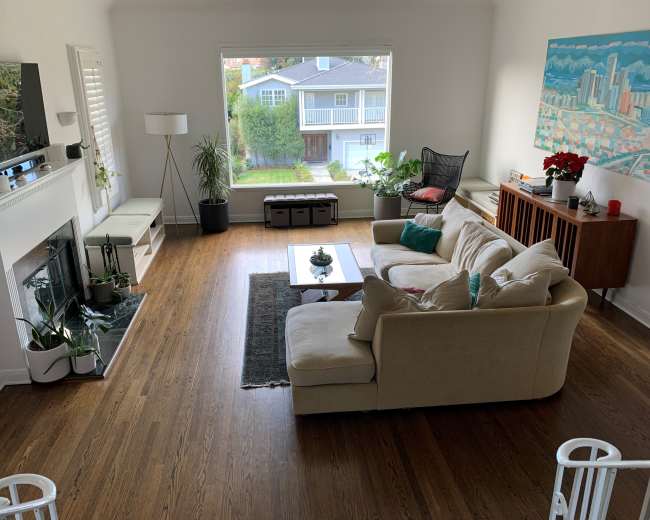 A spacious living room features a beige sofa, a coffee table, a flat-screen TV mounted above a fireplace, and a large window overlooking a garden area.