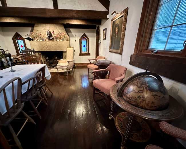 The image shows a room with a wooden dining table, a globe on a stand, and a fireplace, accented by Gothic-style windows and a portrait on the wall.
