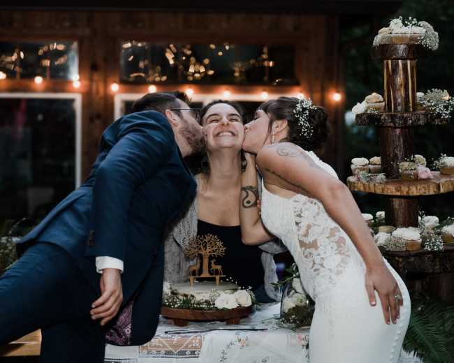 A bride and groom kiss a woman between them at a wedding reception, surrounded by a decorated table and cupcakes.