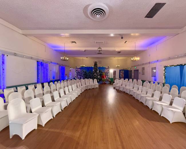 The image shows a spacious indoor venue arranged with white chairs in a circular formation, decorated with blue lighting, and featuring a Christmas tree at one end.