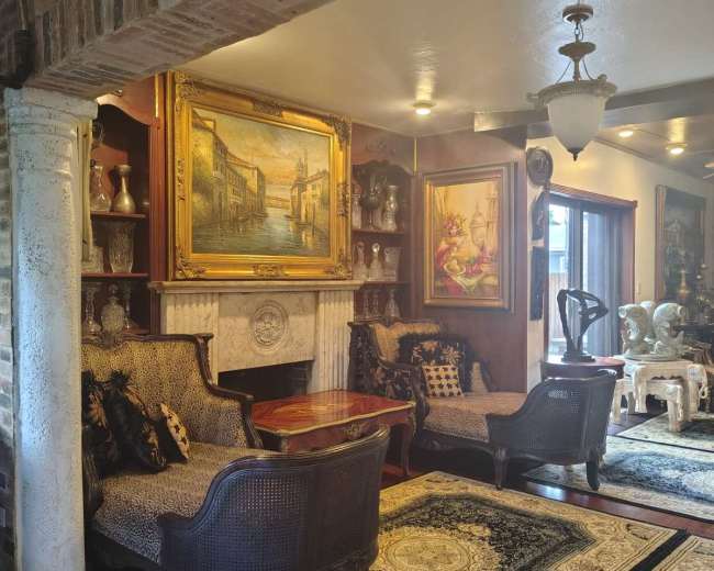 A cozy living room features ornate furniture, a fireplace with a marble mantle, and art framed on the walls.
