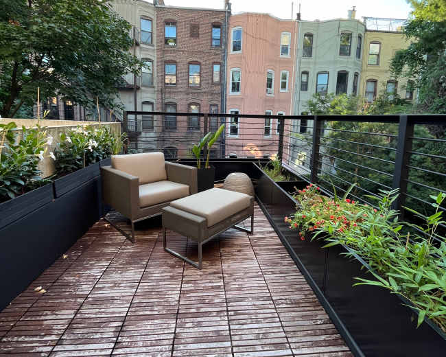 The image shows a small rooftop terrace with two chairs, a small table, and various potted plants, overlooking a row of colorful buildings.