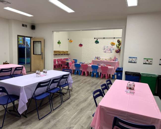 A bright room set up with pink and blue tables and chairs, featuring a play area visible through an open door.