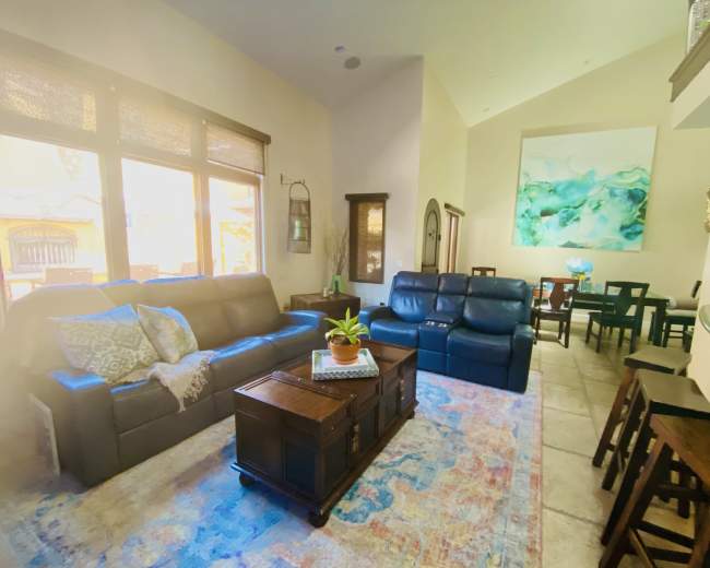 The image shows a bright living room with two sofas, a coffee table, and a dining area in the background.