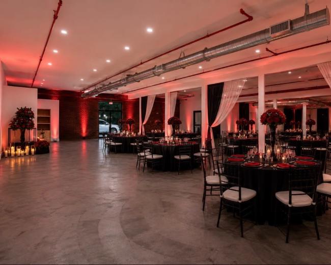 The image depicts a spacious, elegantly arranged event hall featuring round tables set with black tablecloths, red accents, and tall floral centerpieces, illuminated by soft lighting and candles.