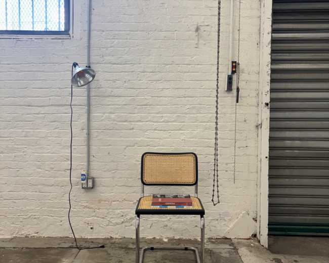 A metal chair with a cane seat is positioned against a white brick wall, illuminated by a lamp hanging nearby.