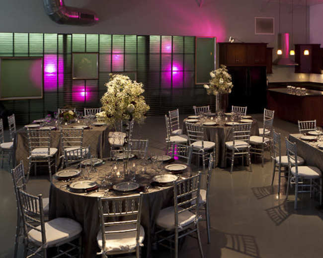 A spacious dining area with several round tables set with elegant tableware and floral centerpieces, illuminated by soft colored lighting.