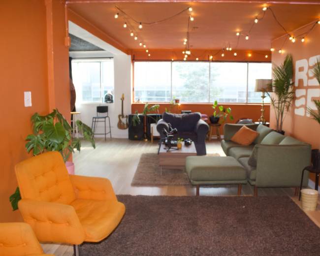 A spacious, well-lit room features orange walls, a mix of seating areas with plants, and string lights overhead.