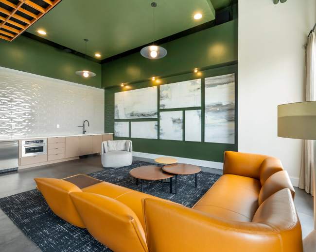 The image shows a modern living room featuring an orange sofa, a circular coffee table, a kitchenette, and green accent walls adorned with abstract artwork.