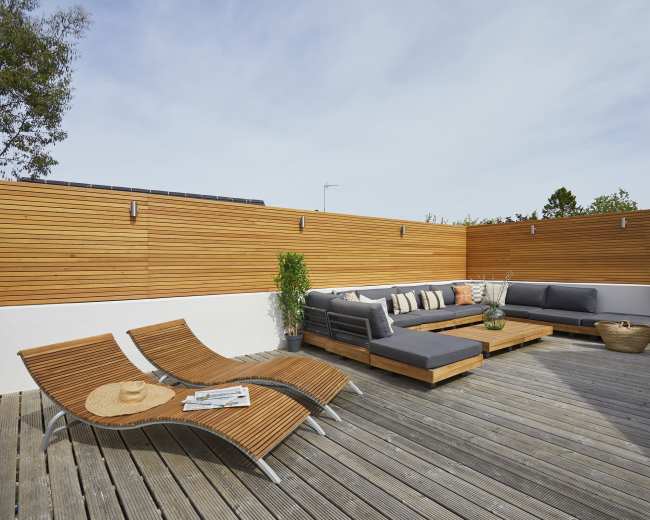 A modern outdoor terrace with a wooden deck featuring a sectional sofa, a coffee table, two lounge chairs, and decorative plants against a wood-paneled wall.
